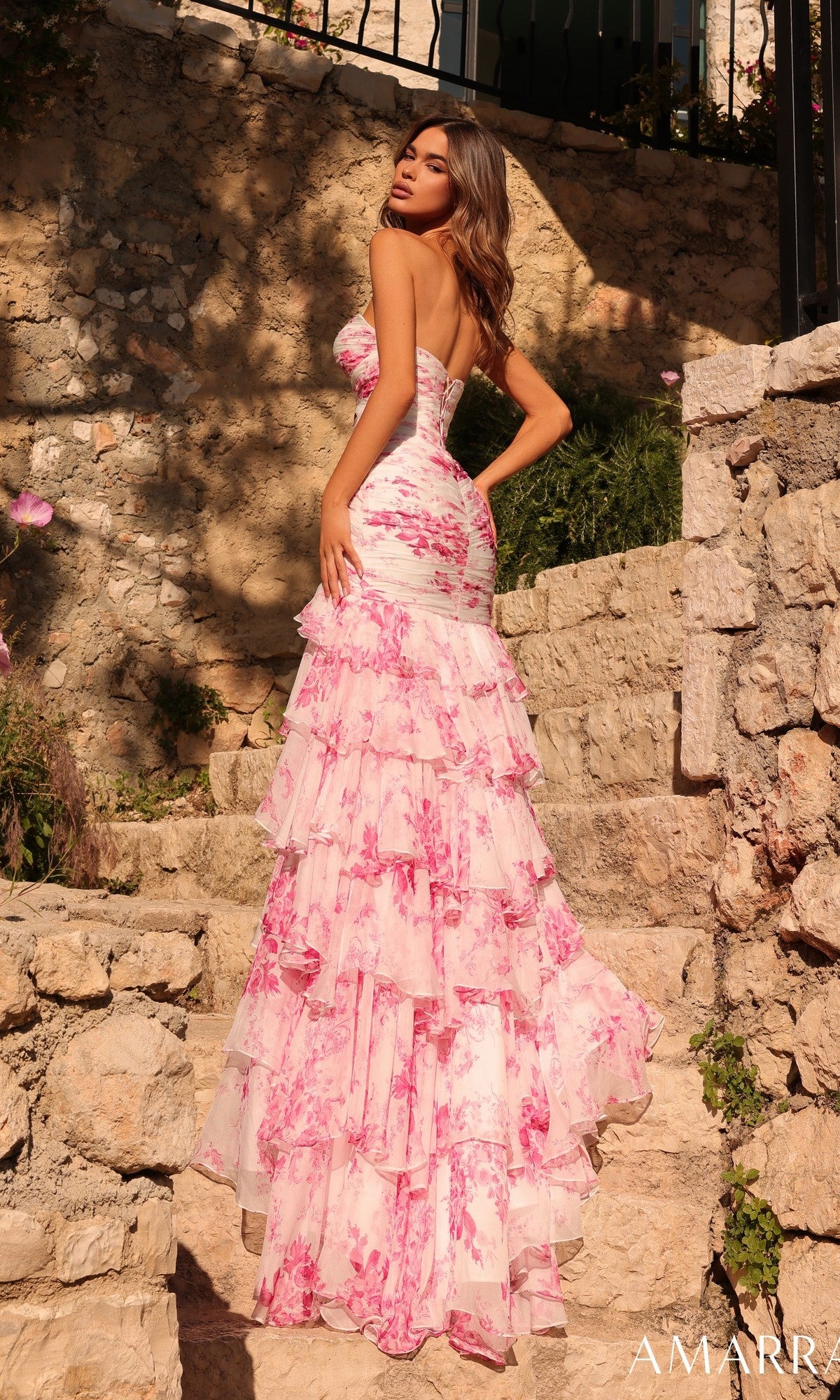 A woman in the Amarra 89297 long prom dress—strapless chiffon with pink floral ruffles—stands on stone steps outdoors, turned slightly away from the camera.